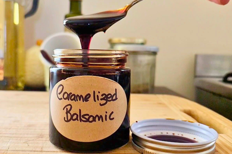 A jar of thick, glossy homemade caramelised balsamic glaze on a wooden board with a spoon beside it.
