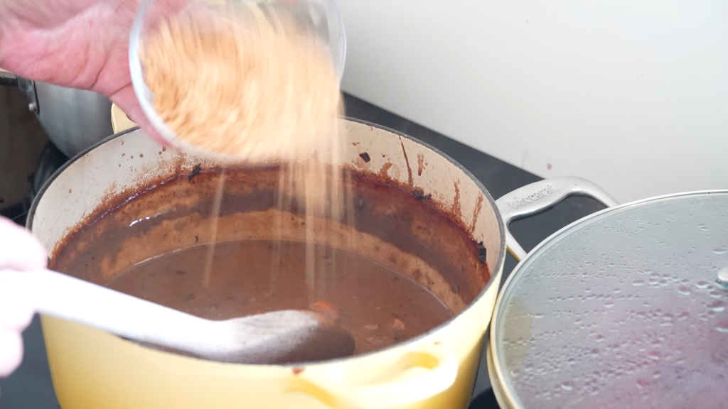 A close-up of ginger nut biscuit crumbs being added to a rich brown sauce in a yellow pot