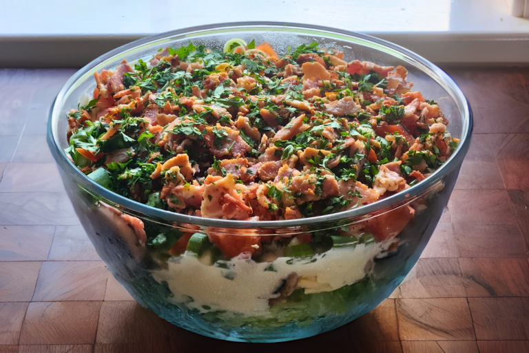 Glass bowl layered salad with chopped bacon, peas, eggs, cheese, and fresh parsley.