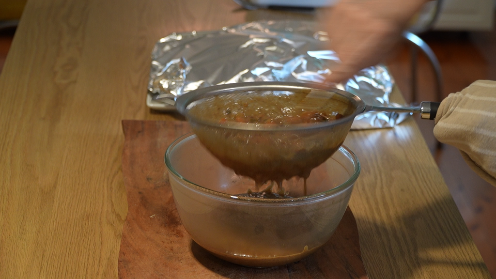 Thickened Sauerbraten sauce being strained into a glass bowl through a metal sieve