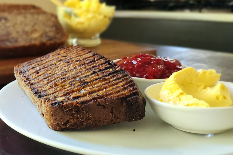 A slice of toasted banana bread served with honey butter and raspberry jam on a white plate.