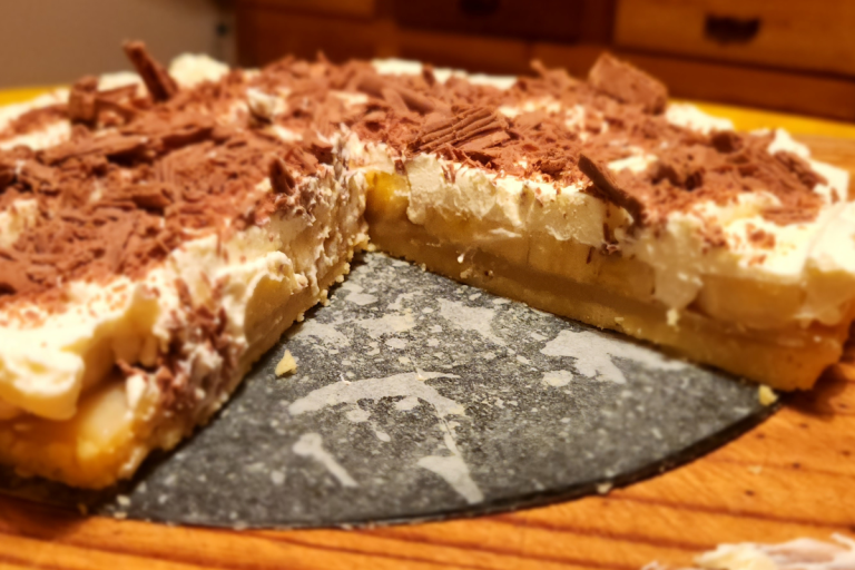 Banoffee Pie in a tart tin with a slice removed, showing layers of banana, caramel, and whipped cream