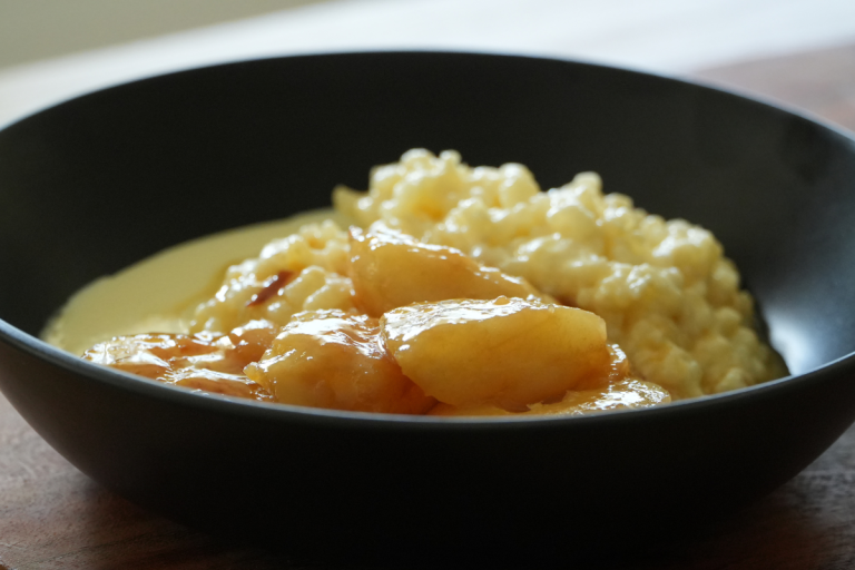 A close-up of creamy oven-baked rice pudding served with golden caramelised pears in a black bowl.