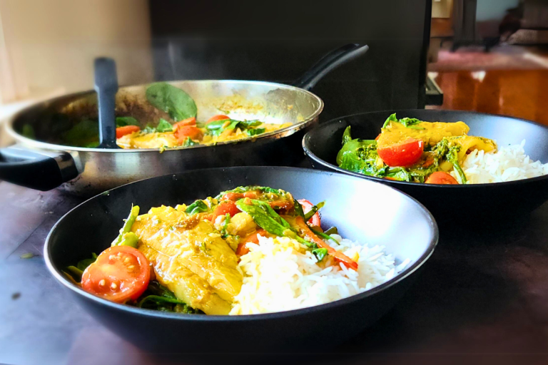 Two steaming black bowls of fish curry with rice, spinach, cherry tomatoes, and capsicum.