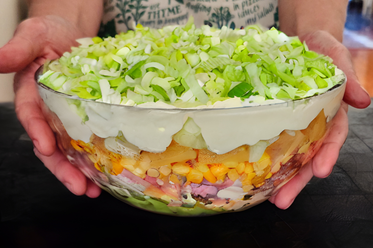 Layered German salad in a glass bowl with leeks, pineapple, corn, ham, and creamy dressing, held in hands