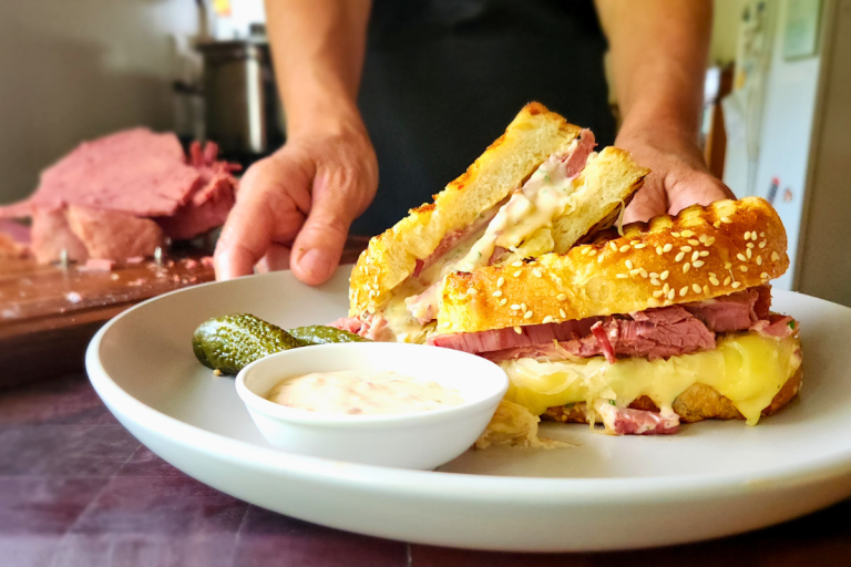 Homemade Reuben sandwich with melted cheese, corned beef, and pickles on a white plate.