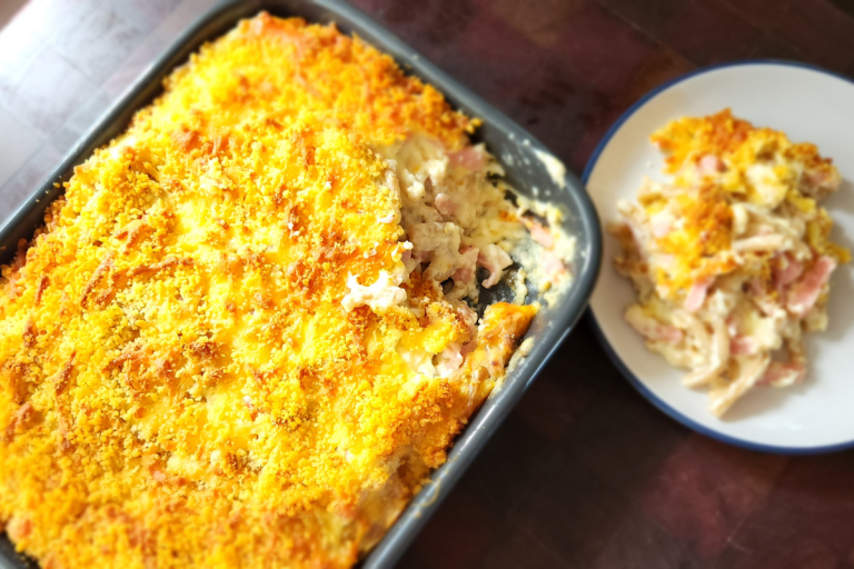 A baked dish of creamy ham macaroni and cheese with a golden breadcrumb topping, served with a portion on a white plate.
