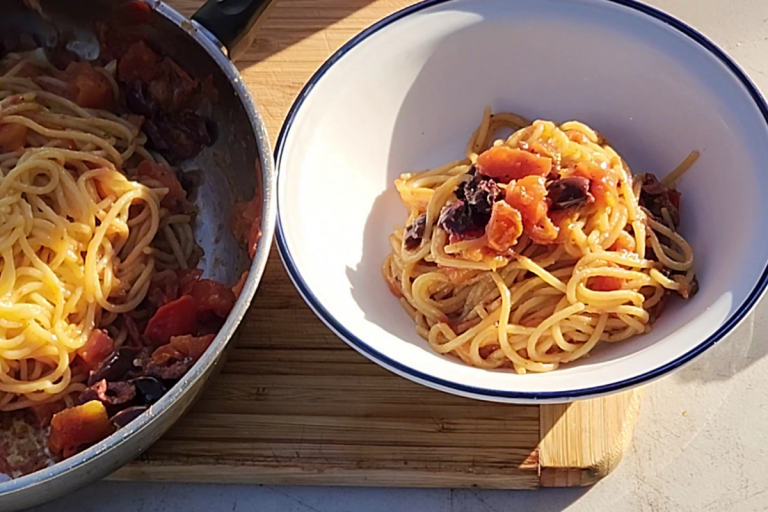 A bowl of anchovy pasta next to a pan of spaghetti with tomatoes and olives