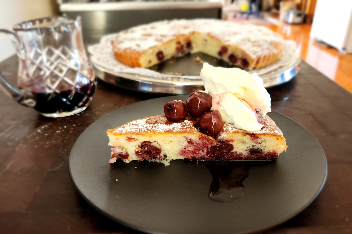 Slice of cherry cake with syrup jug, whipped cream, and the full cake in the background.