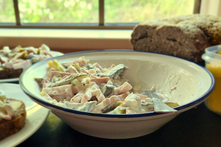 German Fleischsalat in a serving bowl with a spoon, fresh bread and butter in the background