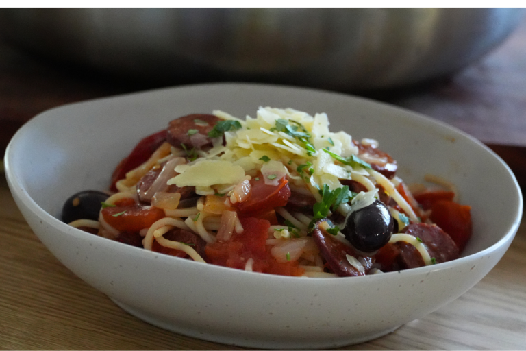 Close-up of chorizo pasta topped with shaved cheese, olives, and fresh parsley in a white bowl.