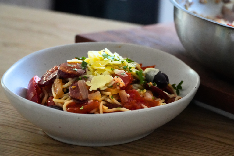 Bowl of easy chorizo pasta served with olives, tomato, and fresh parsley, sitting on a timber benchtop.