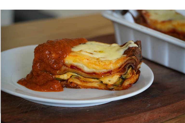 Slice of vegetarian lasagne topped with béchamel and extra tomato sauce on a white plate, with the baking dish in the background.