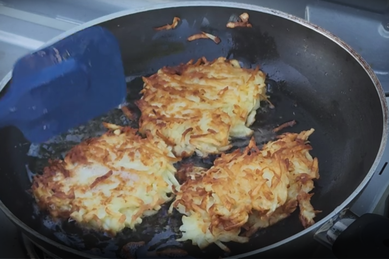 Three crispy potato pancakes frying in a black pan over a camp stove, turned with a blue spatula.
