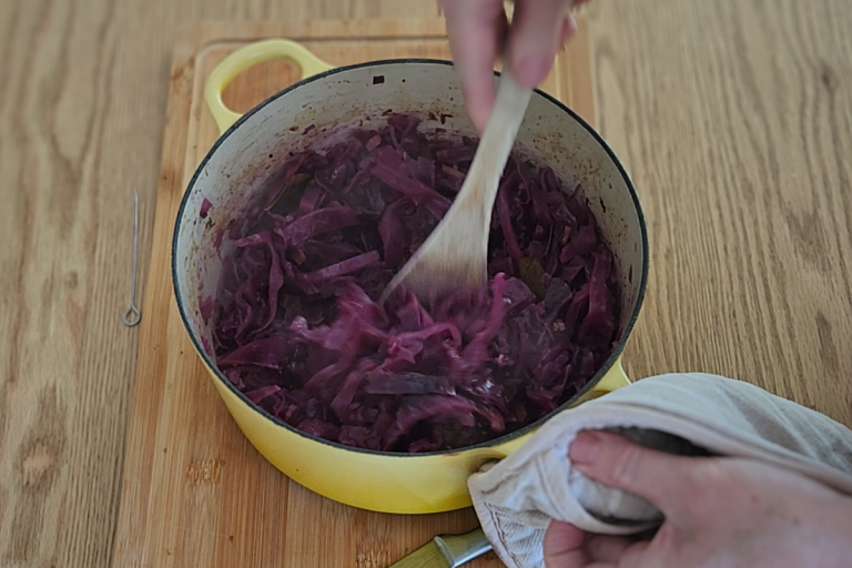Braised red cabbage in a yellow Dutch oven being stirred with a wooden spoon.