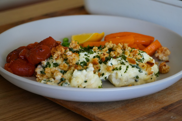 Close-up of homemade fish pie with creamy mashed potatoes, parsley, and golden breadcrumbs, served with sautéed tomatoes, peas, and honeyed carrots.