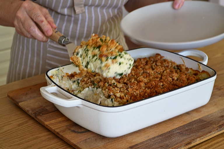 A spoonful of creamy homemade fish pie with a golden breadcrumb topping being served from a white baking dish.