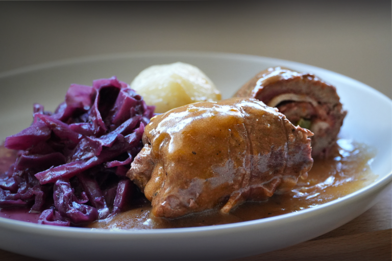 German Rouladen served with red cabbage, potatoes and a rich brown gravy on a white plate.