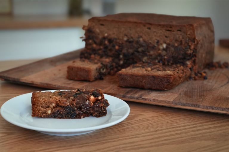 Slice of rich 100-year-old fruit cake on a white plate with the whole cake in the background.