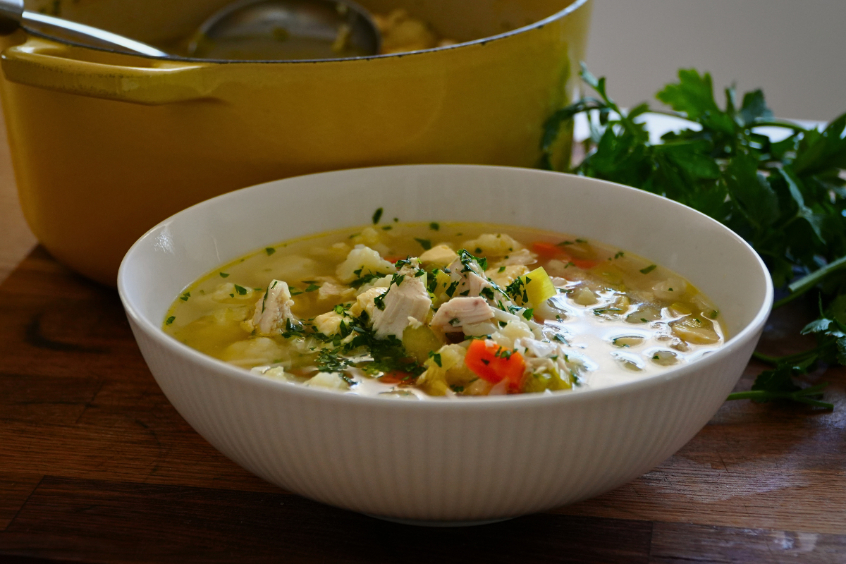 Clear chicken soup with tender chicken, vegetables and parsley in a white bowl with a pot in the background