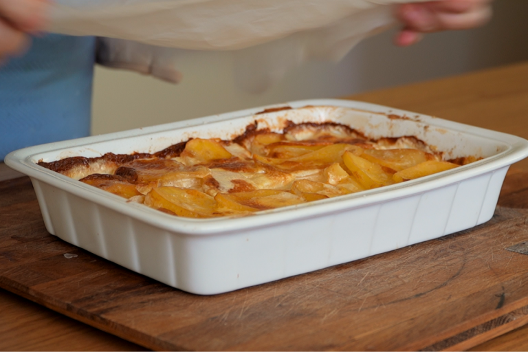 Fresh from oven creamy potato bake being uncovered in white baking dish for crumbed chops recipe