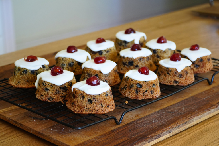 Homemade little fruit cakes cooling on a rack with white icing and cherries on top