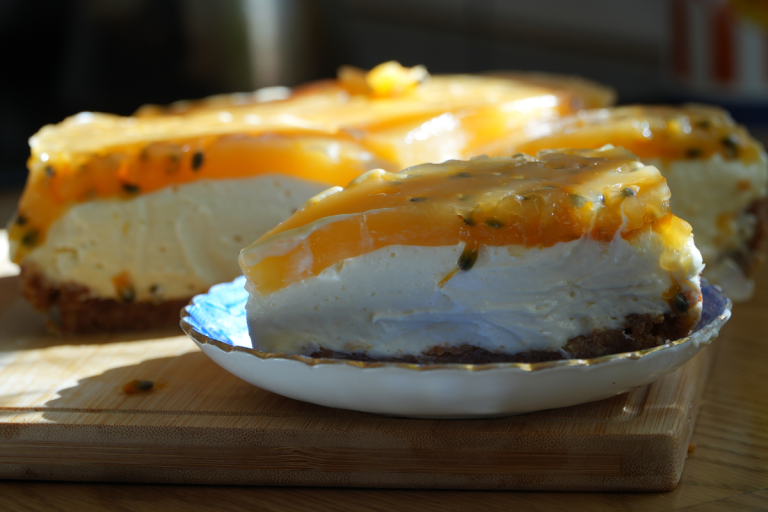 Close-up slice of creamy no bake cheesecake with passionfruit topping on a small plate with more cheesecake in the background.