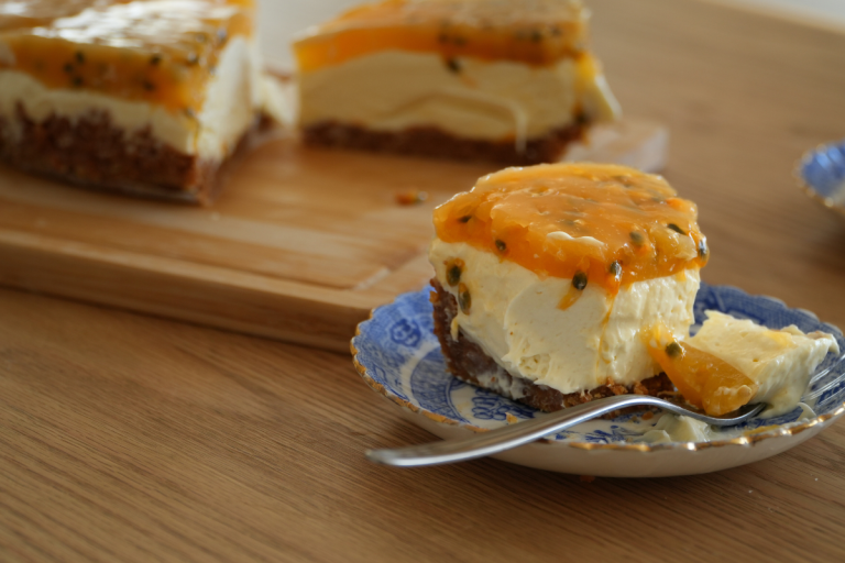 Slice of no bake cheesecake with passionfruit topping on a blue plate with more cheesecake in the background.
