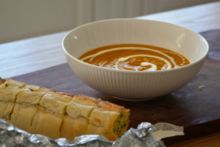Homemade roasted pumpkin soup in a white bowl with roasted garlic bread on a wooden board.